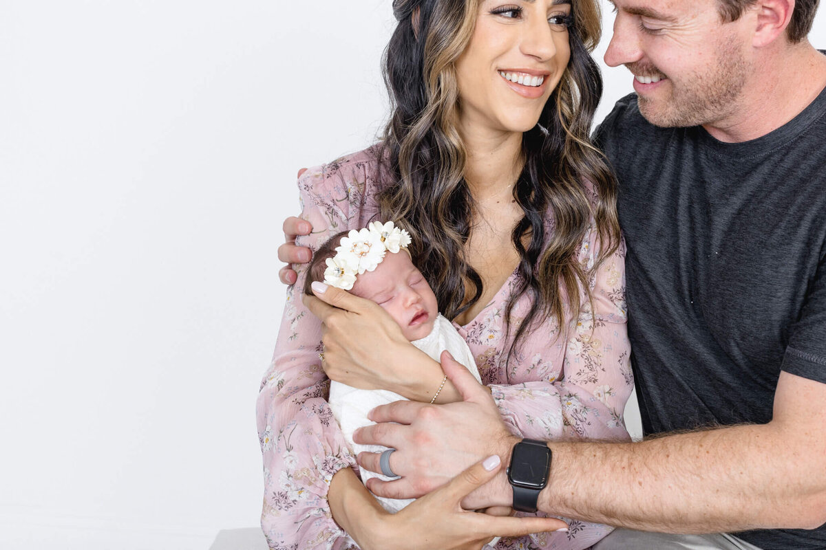 A couple sits closely together, smiling warmly at each other, while holding a sleeping newborn baby. The baby is wrapped in a white blanket and wears a floral headband. The background is plain white.
