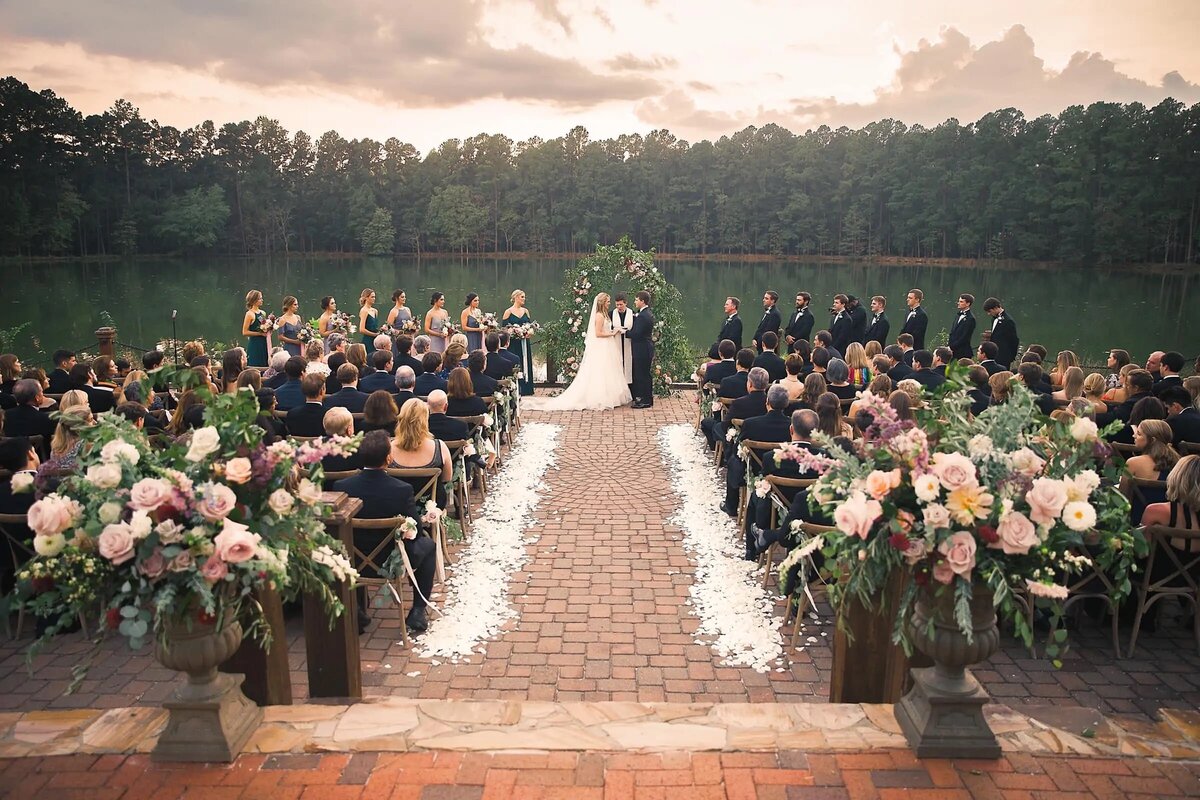An outdoor wedding ceremony set against a tranquil lake, with guests seated on either side of a flower-lined aisle