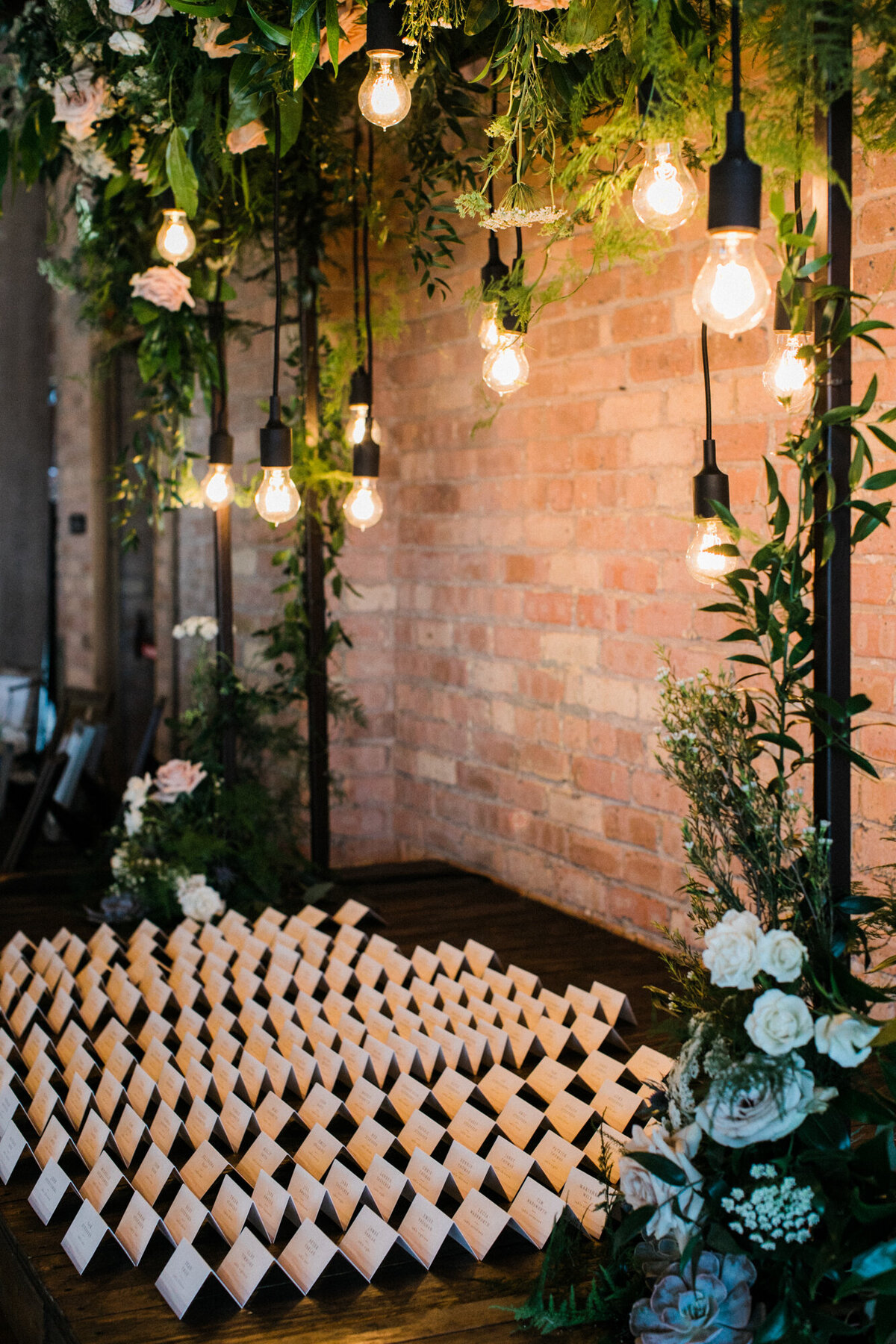 A stylish wedding escort card display, illuminated by warm Edison bulbs.
