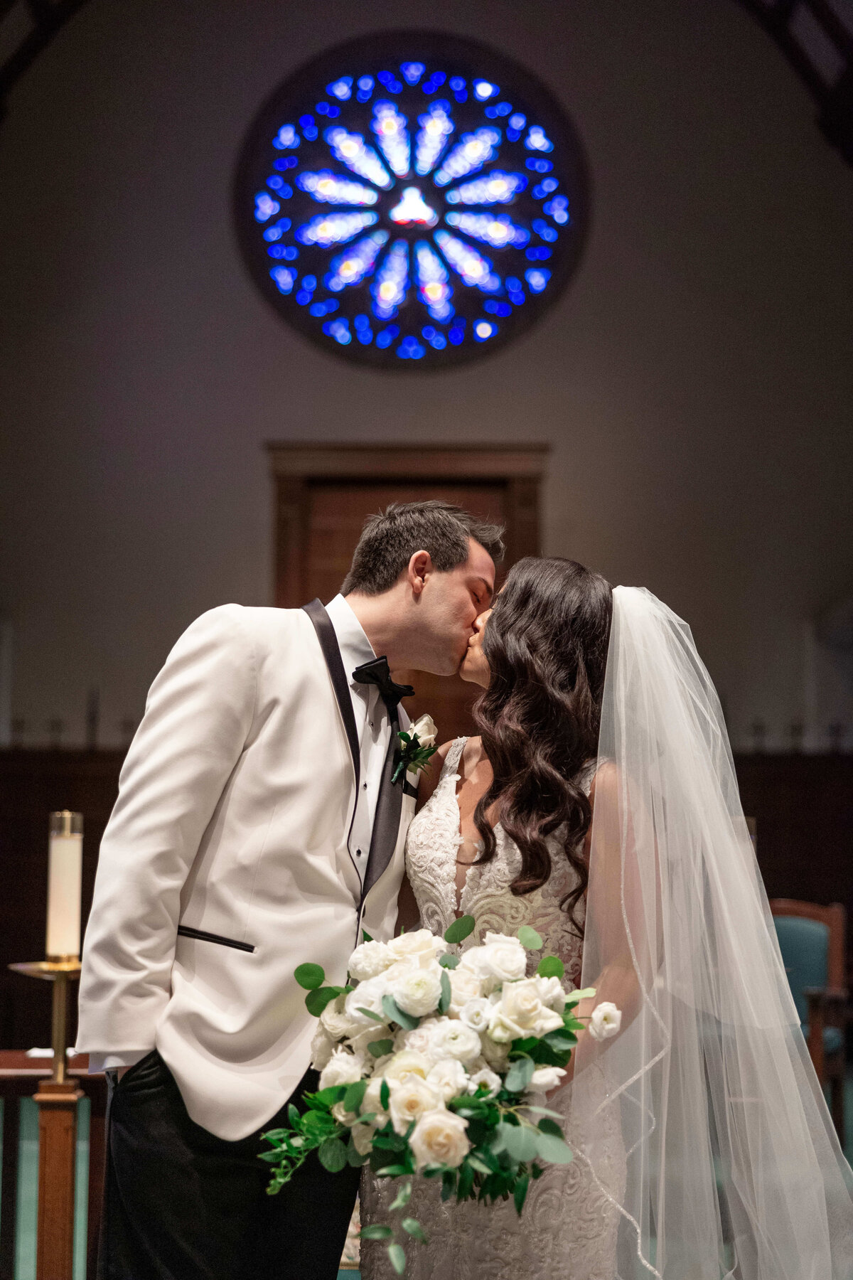 A couple shares a kiss on their wedding day, standing in a church. The groom wears a white tuxedo and the bride is in a lace gown with a veil, holding a bouquet of white roses. A large, circular stained glass window is visible in the background.