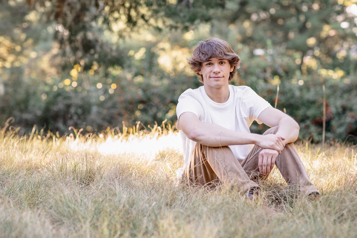 A young man with brunette hair sitting in a grassy field with trees in the background, wearing brown pants and a white shirt, smiling softly at the camera.
