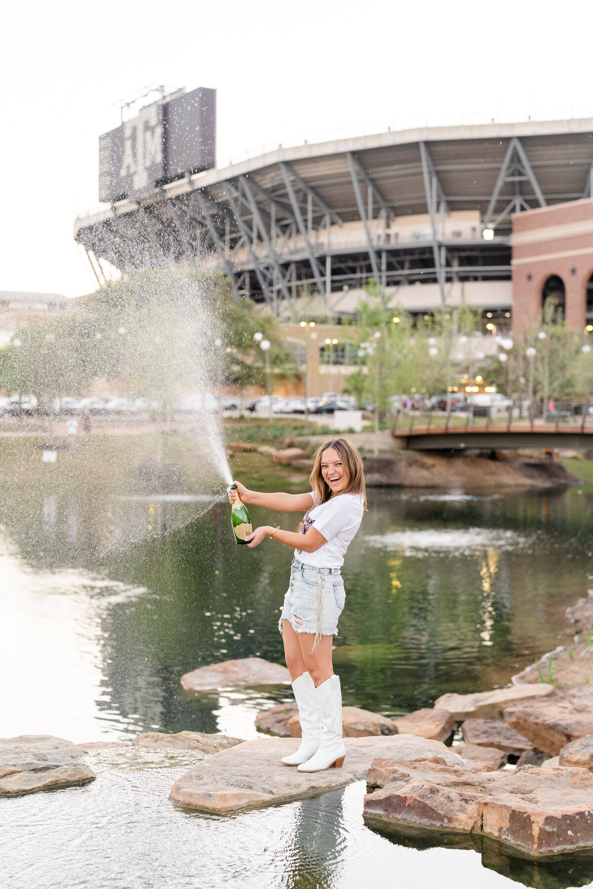 Texas A&M senior girl spraying champagne and laughing while on the rocks of Aggie Park in front of Kyle Field