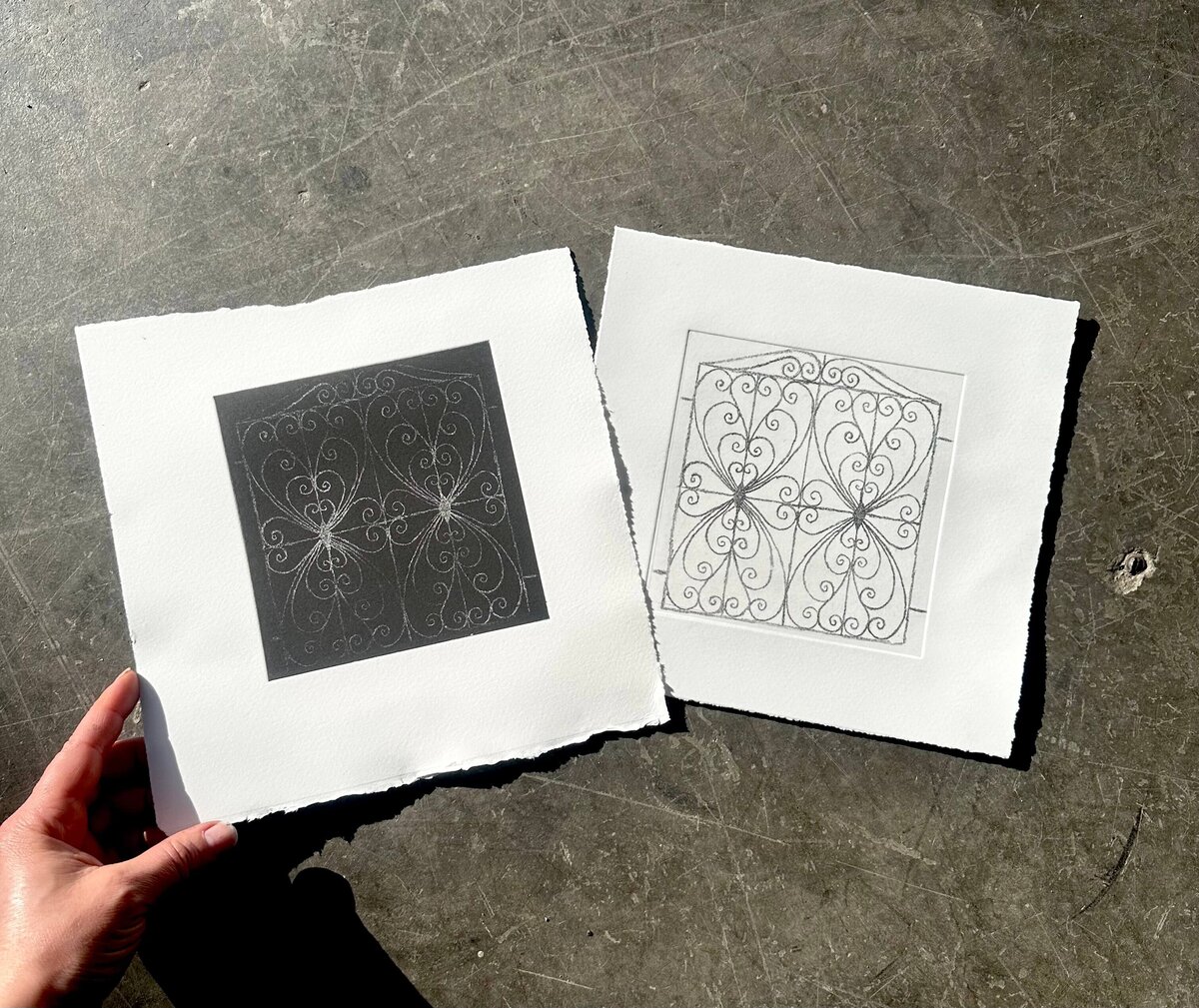 A hand holds two completed prints—one in black, one in white—side by side on dark surface, highlighting contrast and craft.