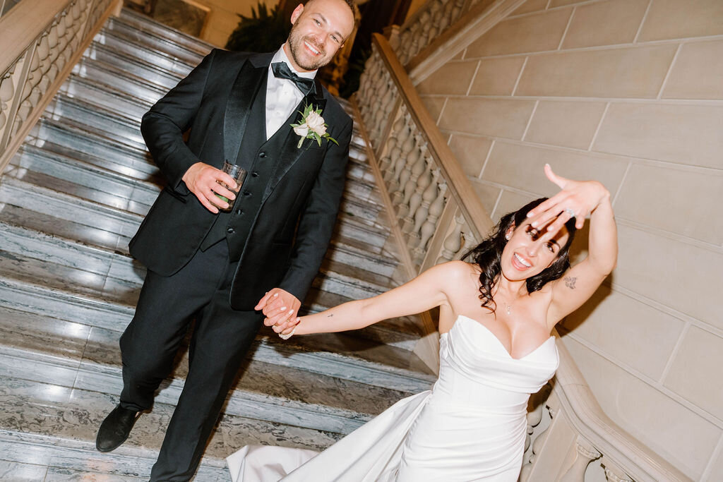 Rhode Island Wedding Photographers | A joyful bride in a white dress playfully gestures while holding hands with the groom in a black tuxedo. They stand on elegant stone steps.