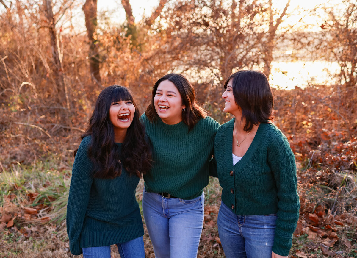 three sisters laughing together while hugging each other and wearing green sweaters at sunset