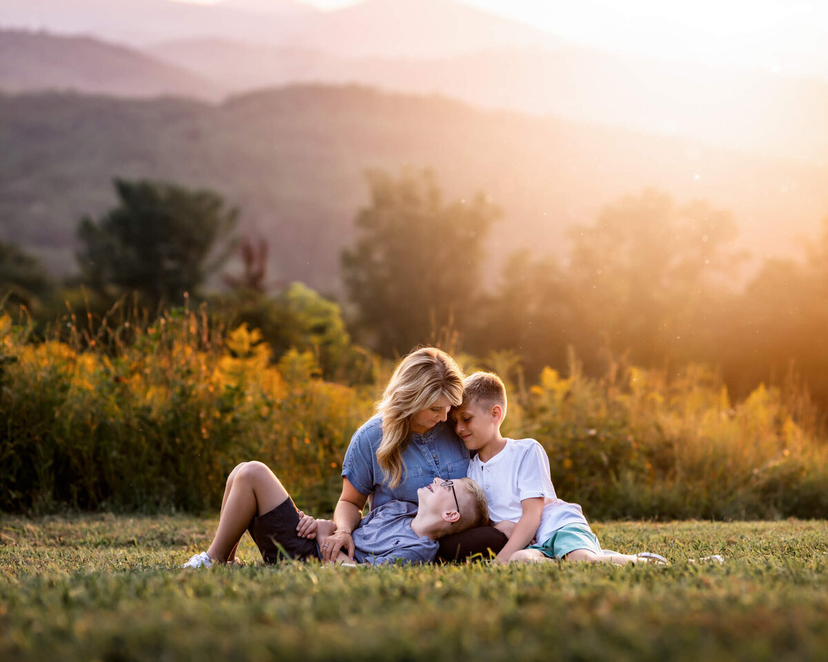 A mom and her sons cuddle and laugh while sitting in the grass with mountains in the distance