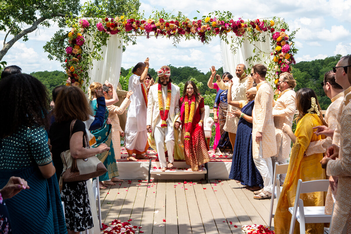 A newlywed couple walks down the aisle under a floral arch at an outdoor wedding ceremony in Illinois, surrounded by guests in colorful attire.