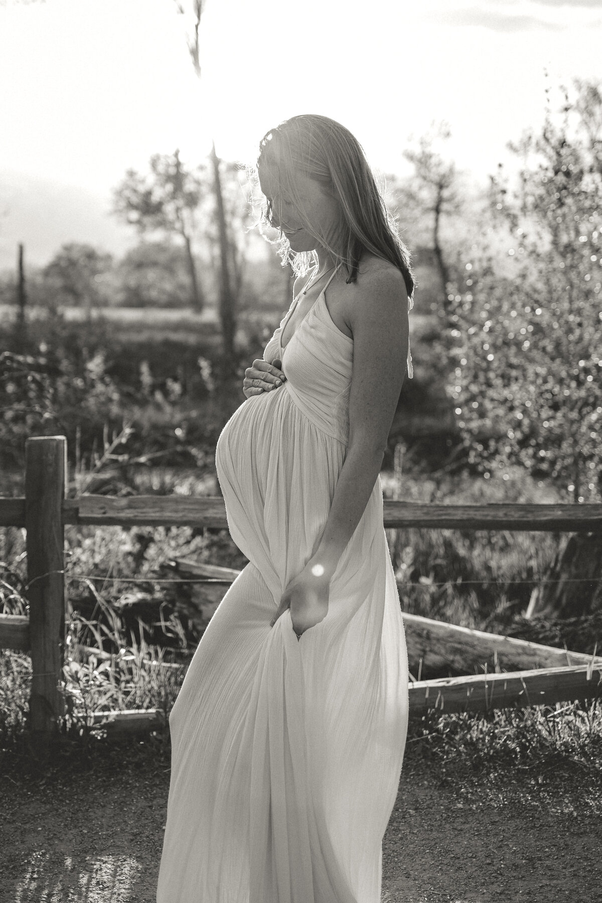 black and white image of expectant mom with hand on top of her belly looking down in white dress with sun flares all around her during maternity session with alexis adkins photography in firestone colorado