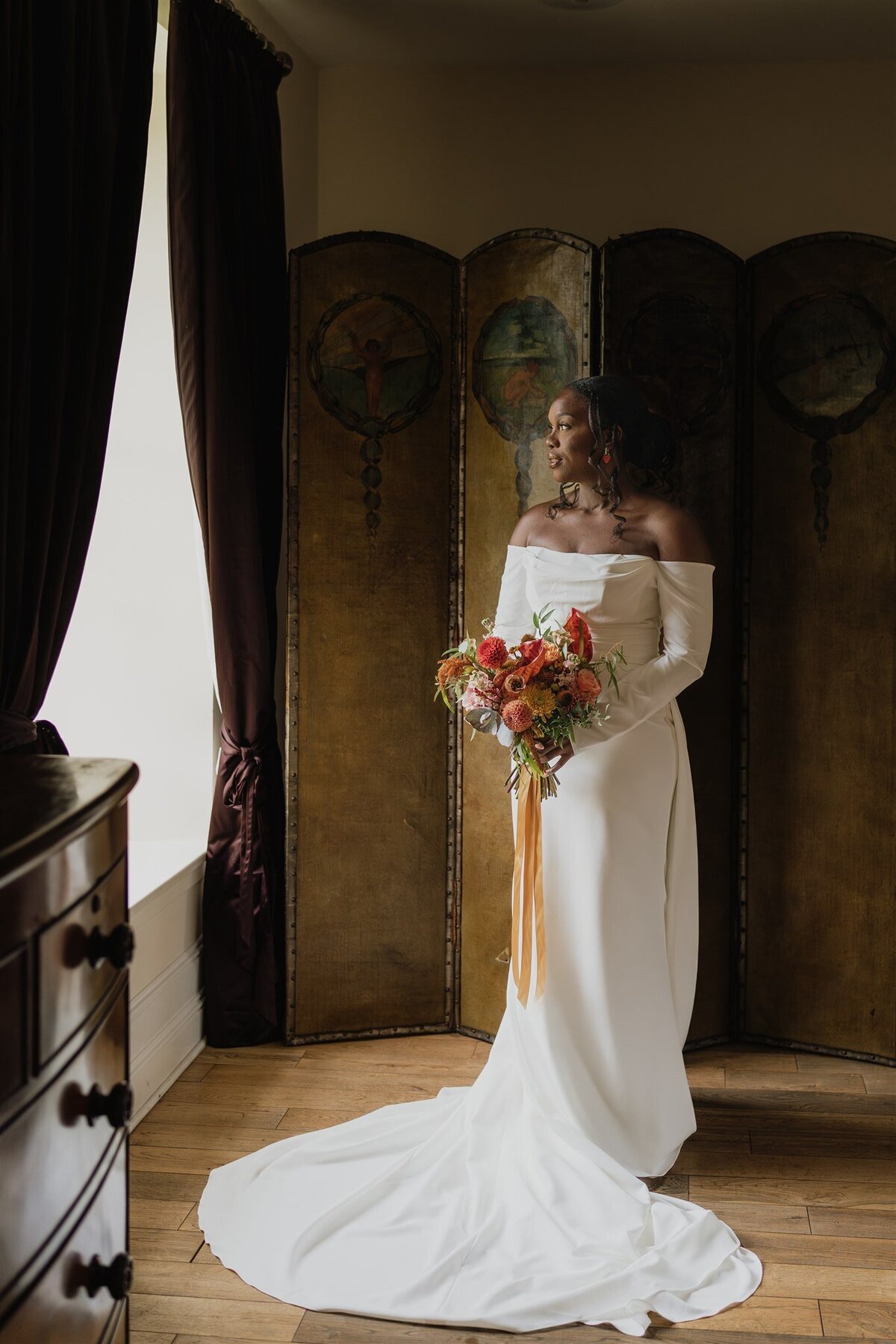 A bride stands wearing her wedding dress and looks out a window towards the Aberdeenshire countryside. She is photographed in front of a traditional wooden privacy screen in the bridal suite of Logie Country House.