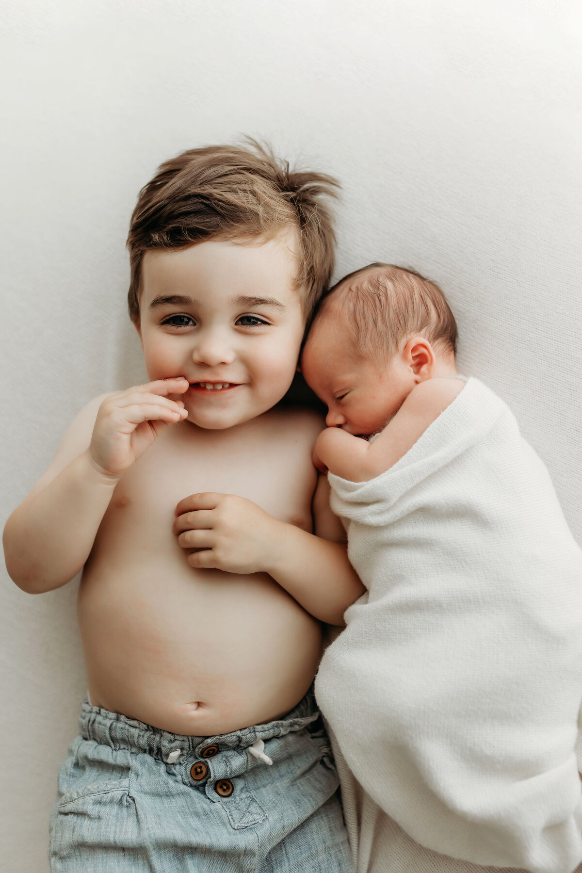 A toddler brother lays happily next to his newborn baby brother