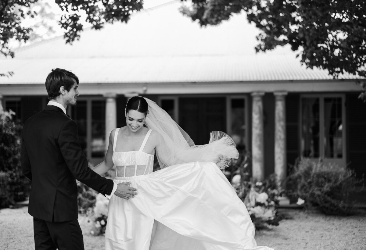A black and white image of a bride and groom on their wedding day. A gush of wind blows the brides dress into the air