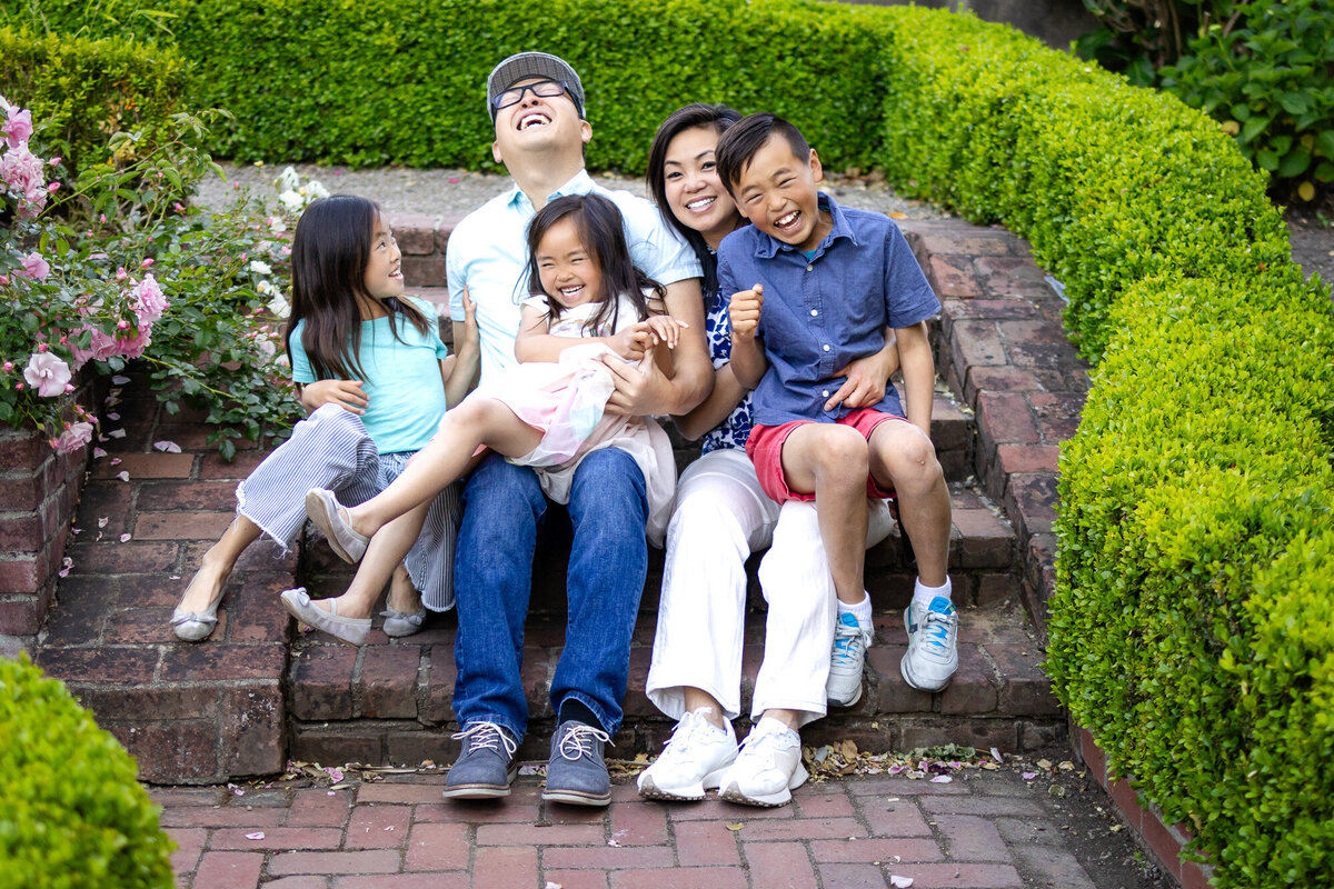 Family of five sitting on garden steps surrounded by lush greenery and blooming roses, sharing a moment of pure laughter — Bay Area Family Portfolio - Ellobelle Photography