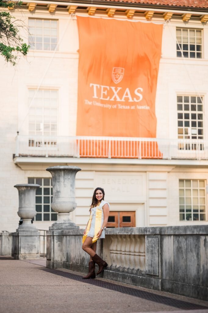 A UT senior in a white dress and brown boots leans against a half wall during her graduation portrait session on campus.