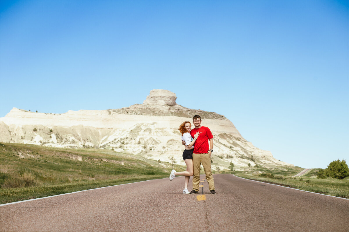 A couple stands on a lined road posing in front of the camera with a large rock mass behind them.