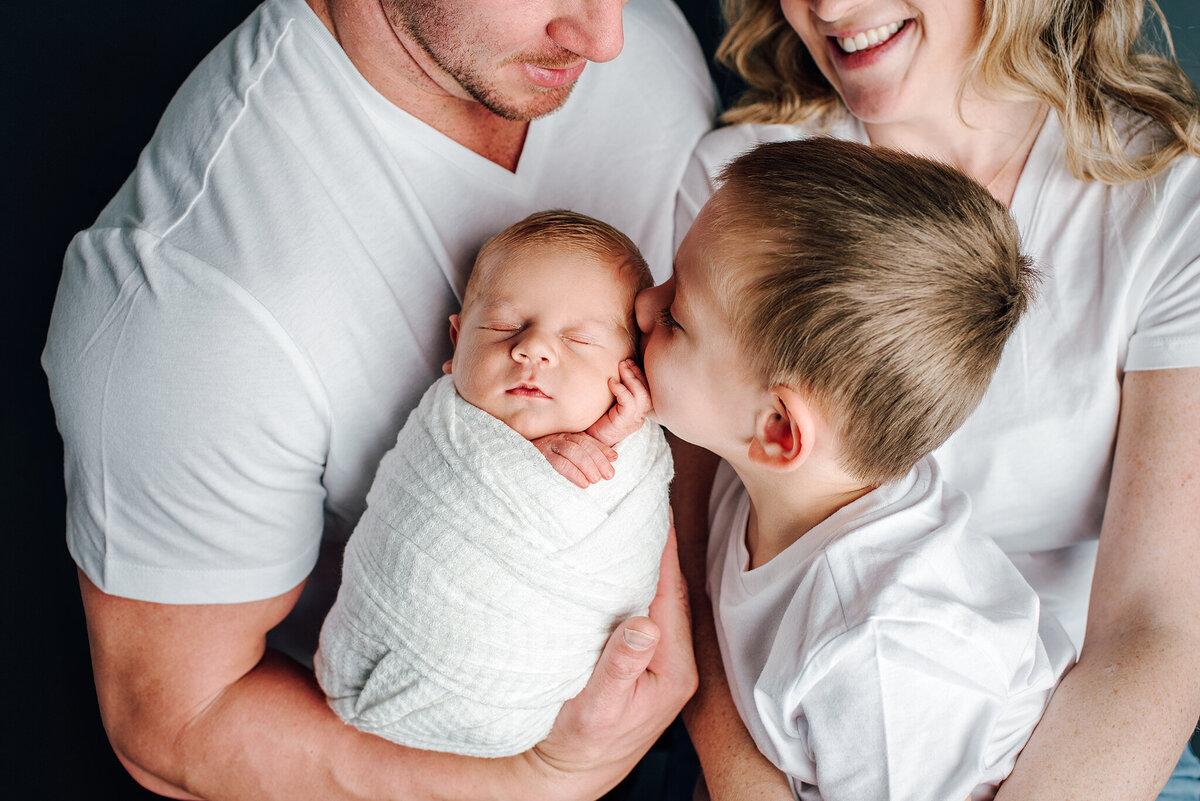 Family of 4 in white shirts holding newborn baby against dark background in Jacksonville, FL.