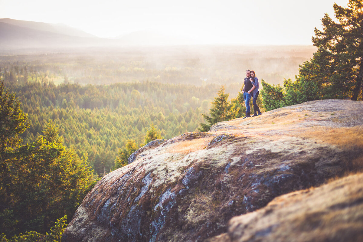 A photo of a couple embracing on a cliff at sunset above a forest.