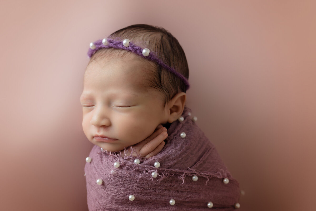Newborn Photographer, a baby girl is swaddled in a purple blanket with pearls sewn on