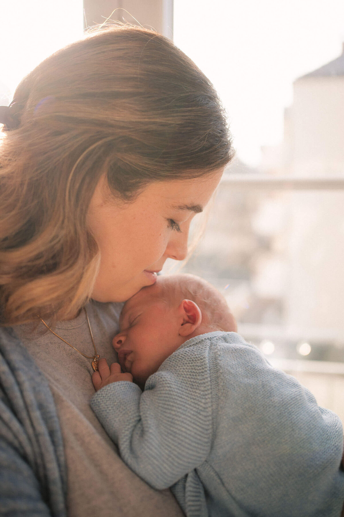 Mother kissing her newborn’s head while holding them close to her chest in soft golden light.