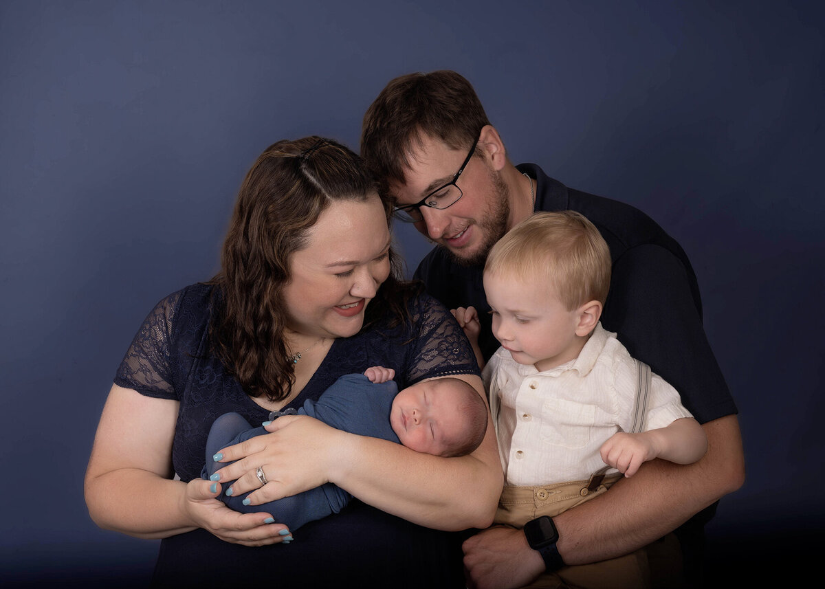Happy parents stand in a studio while introducing their toddler to their newborn baby in mom and dad's arms