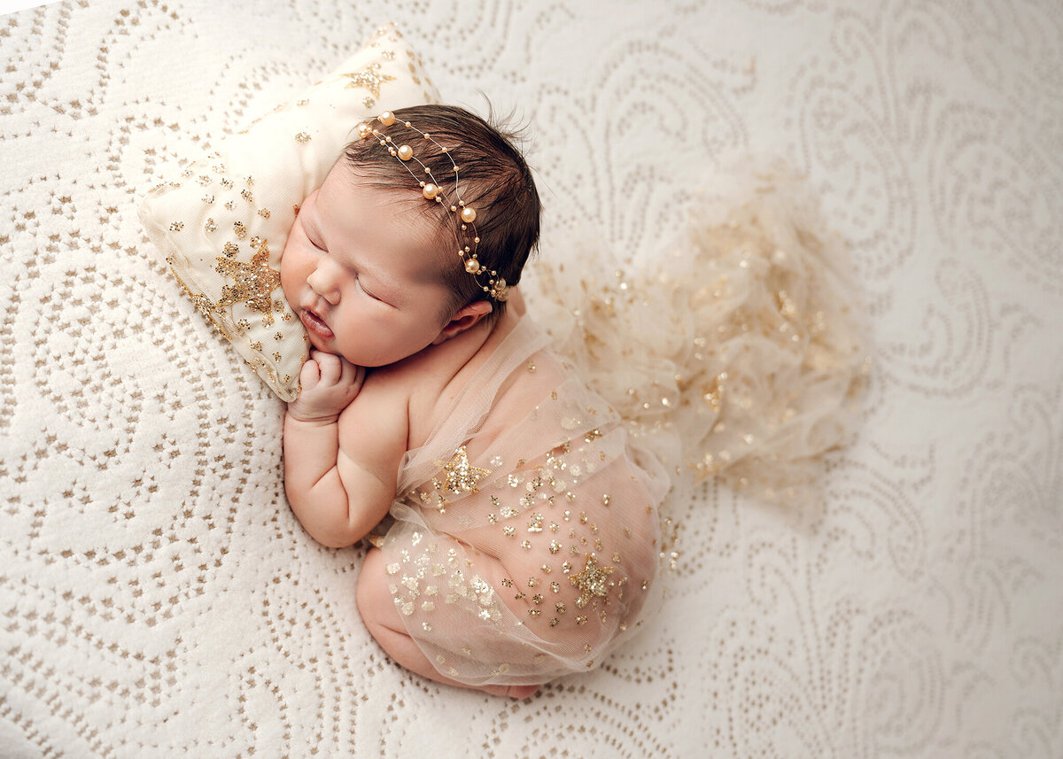 adorable baby girl wrapped with a gold star scarf, with a minimal gold headband on a gold accented pillow on a paisley blanket