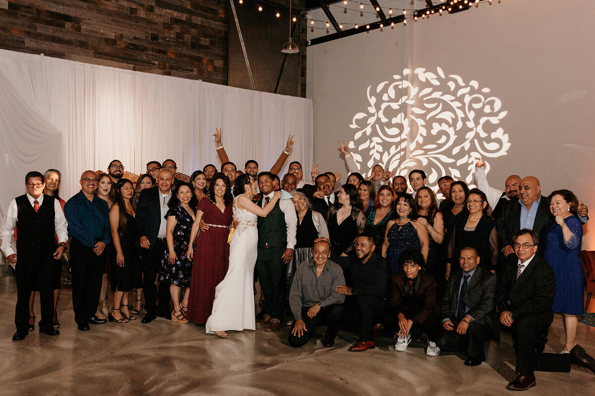 A large group photo of wedding guests posing with the bride and groom at the center, surrounded by friends and family in a warmly lit venue.