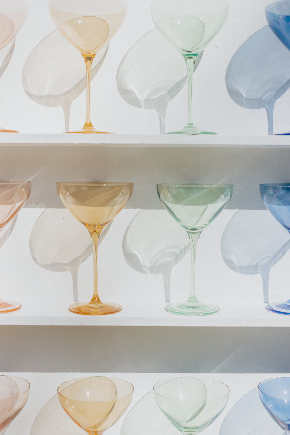 A close-up still life of multicolored cocktail glasses casting beautiful shadows on a white shelf. The composition is airy, modern, and artful, showcasing vintage-inspired glassware in a rainbow palette. Captured by Dani Lacey Photographs in Greenville, South Carolina.