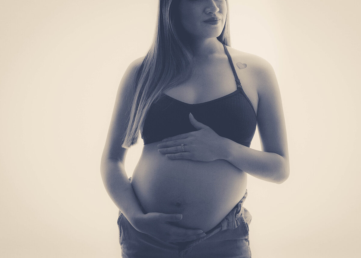 A mom to be stands holding her bump in a white studio in open jeans and black bra