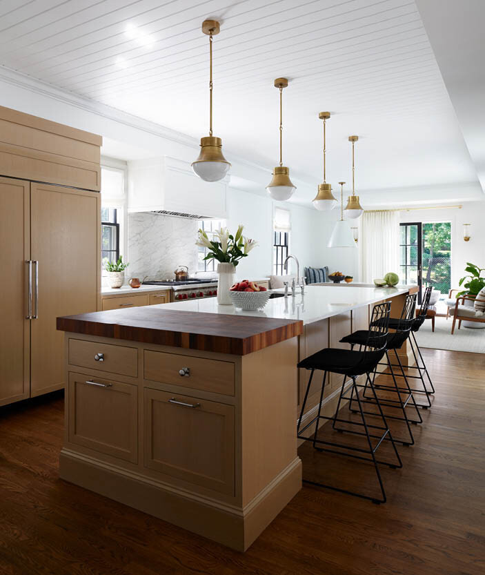 Discover the custom kitchen in a historic Hingham home, featuring oak cabinets, a large island with walnut butcher blocks, brass pendants, and black wire counter stools. No upper cabinets for a unique look.