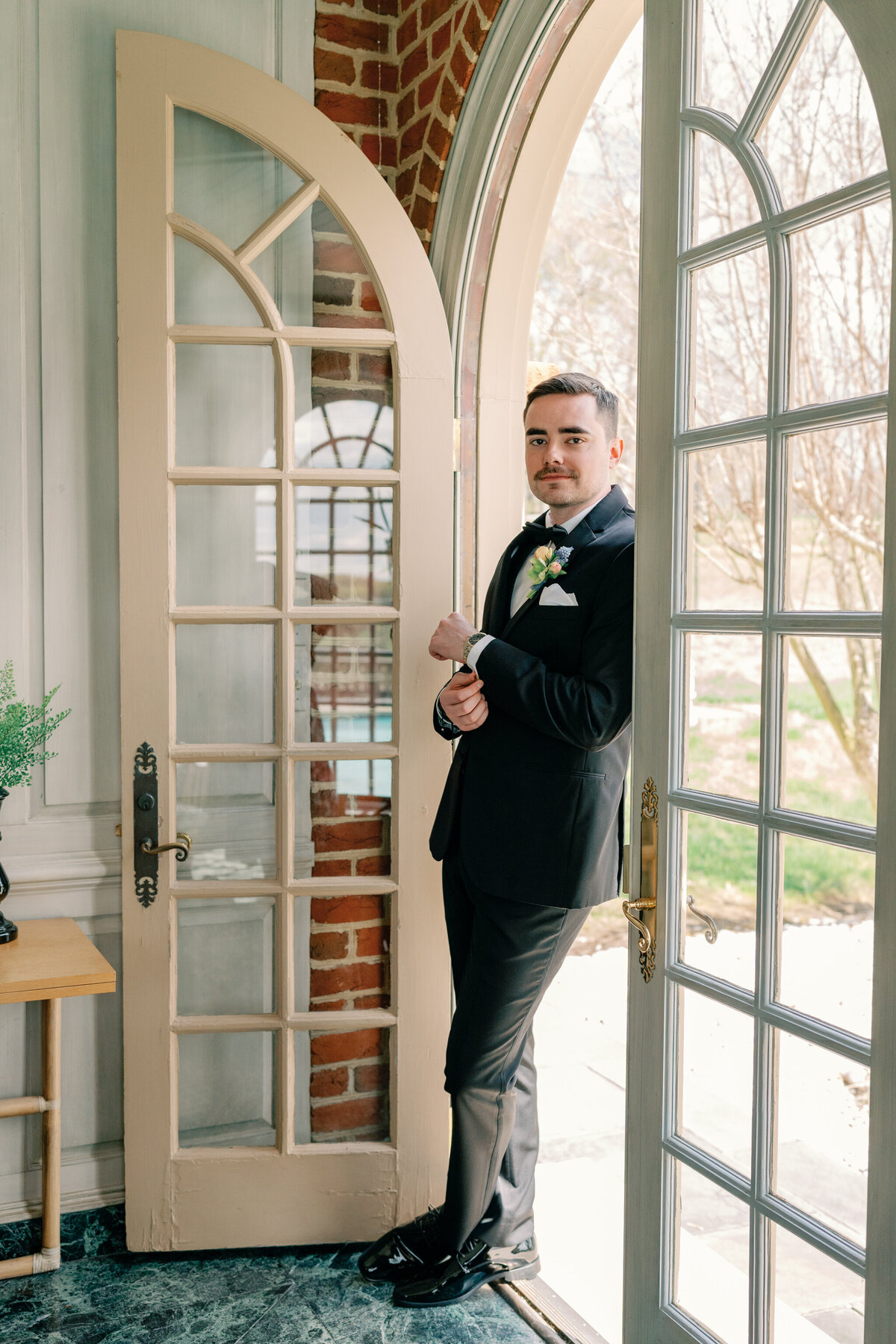Estate at River Run Wedding in Maidens, VA | Wedding Photographer in Richmond | groom in black suit adjusting watch while standing in the doorway of the Estate at River Run pool house getting ready suite