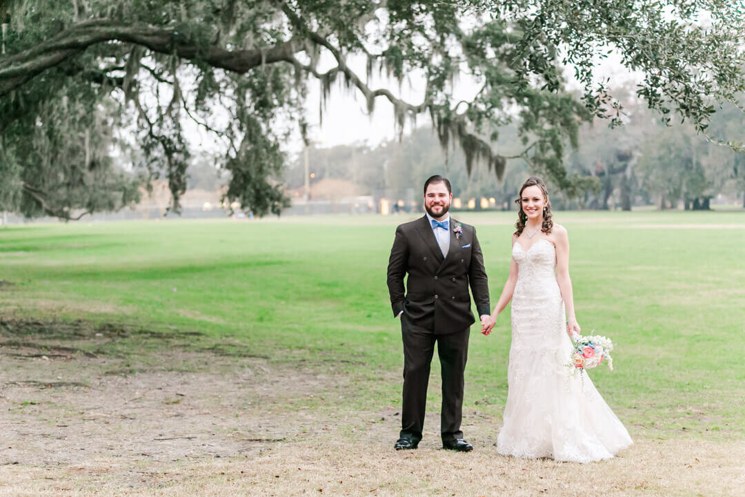 Newlyweds stand in a large lawn under an oak tree holding hands