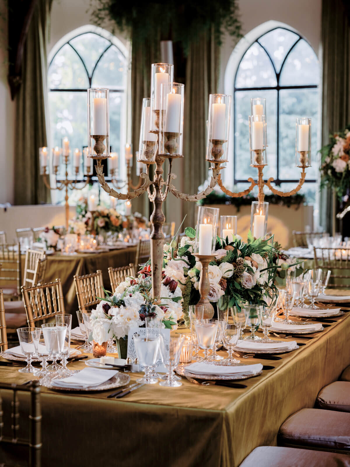 Two long, bronze dining tables and chairs, with large flower bouquet and candle centerpieces, plates and cutleries