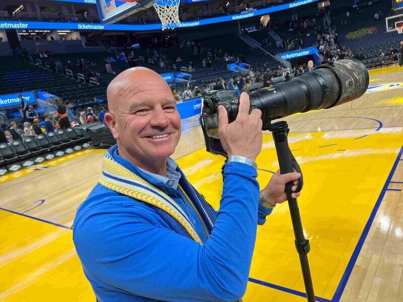 Man smiling on the floor of a basketball game holding a camera with a very long lens