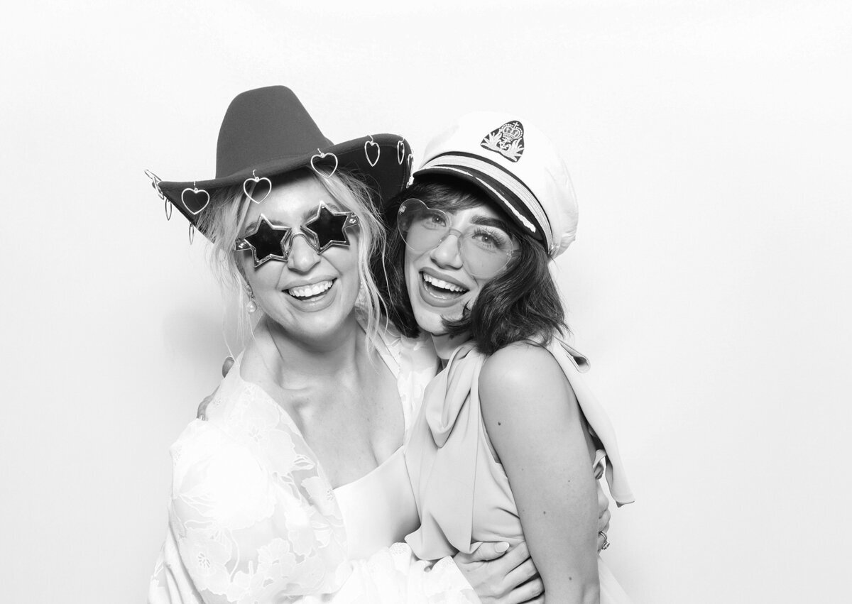 B&W image of 2 guests posing against a white backdrop, with Mad Hat Photo Booth rental for weddings or corporate events, with premium features, for luxury events at Kin House venue