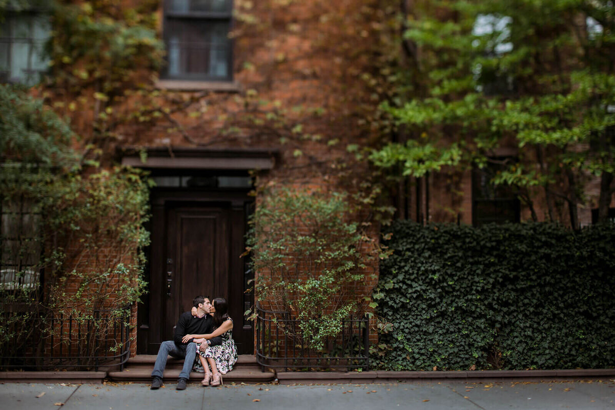 The engaged couple is kissing while sitting in front of an ivy-covered house in West Village, NYC. Image by Jenny Fu Studio.