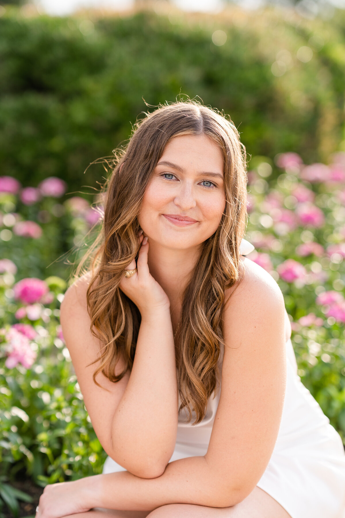 Texas A&M senior girl sitting and leaning on arm with hand on neck and smiling with flowers in the background at the Bell Tower