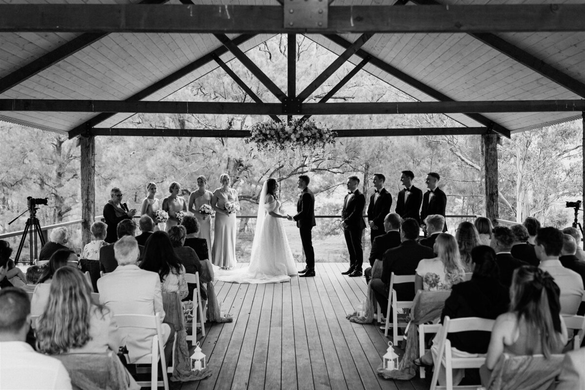 A beaming bride, clutching a bouquet of white roses, exchanges vows with her groom under a canopy of fragrant blossoms.