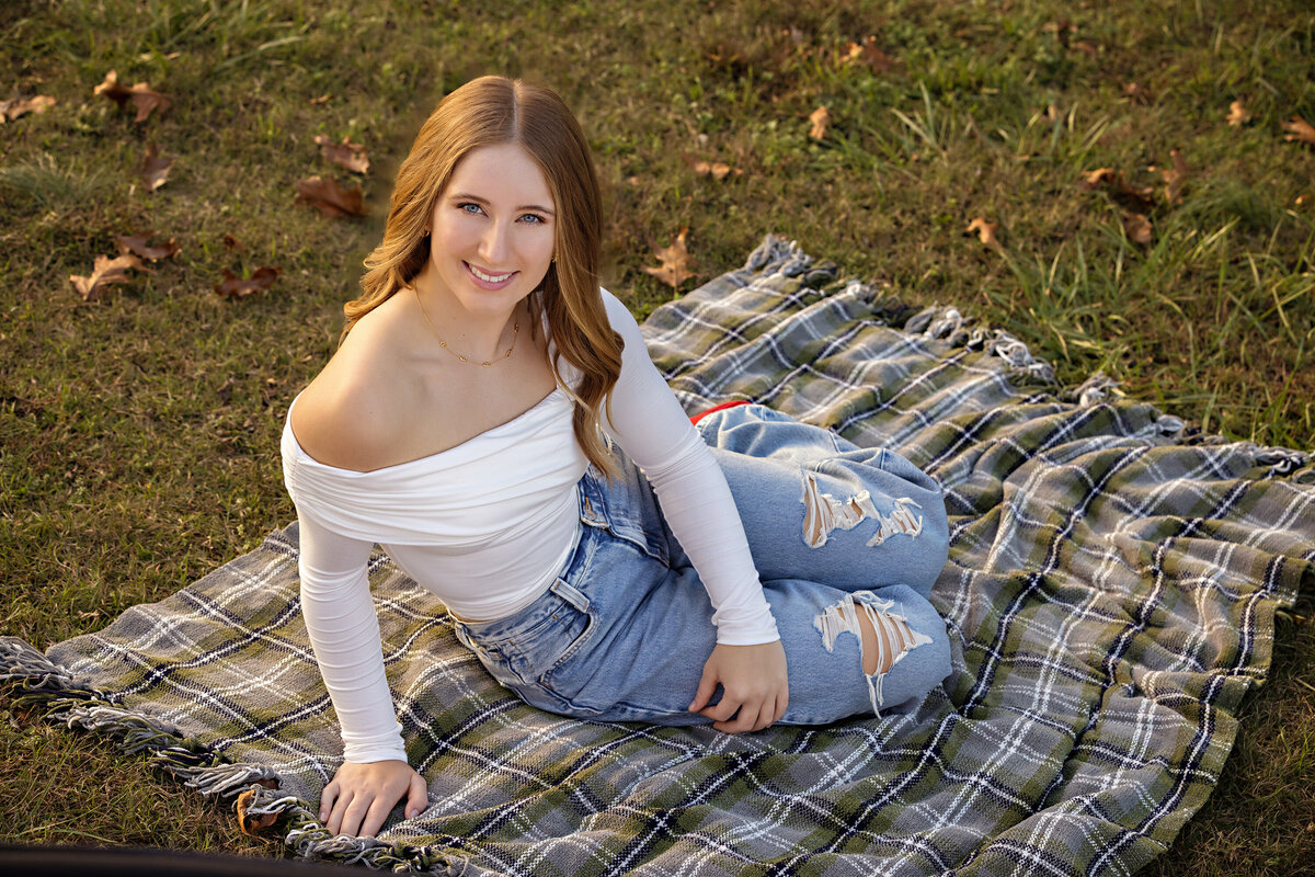 High school senior in jeans and white top sitting on a gray and green and blue plaid blanket on a grassy hillside at Anne Springs Close Greenway