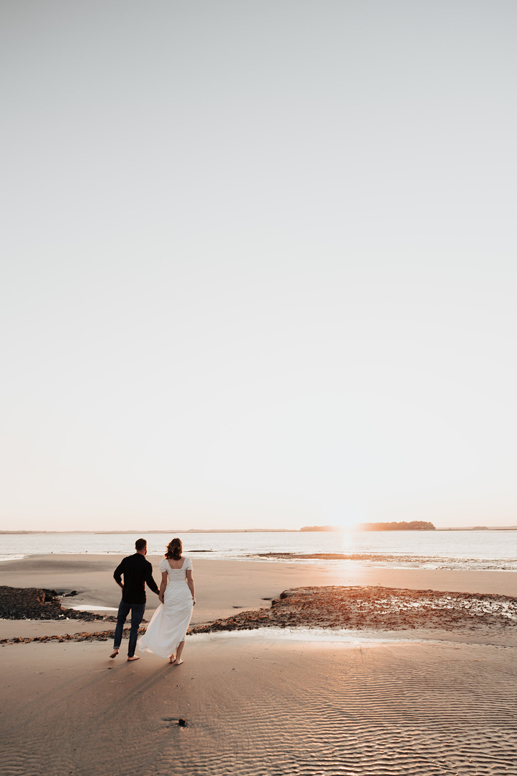 Wide angle photo of engaged couple walking towards ocean. Sun is setting in background. Low tide and low waterline.