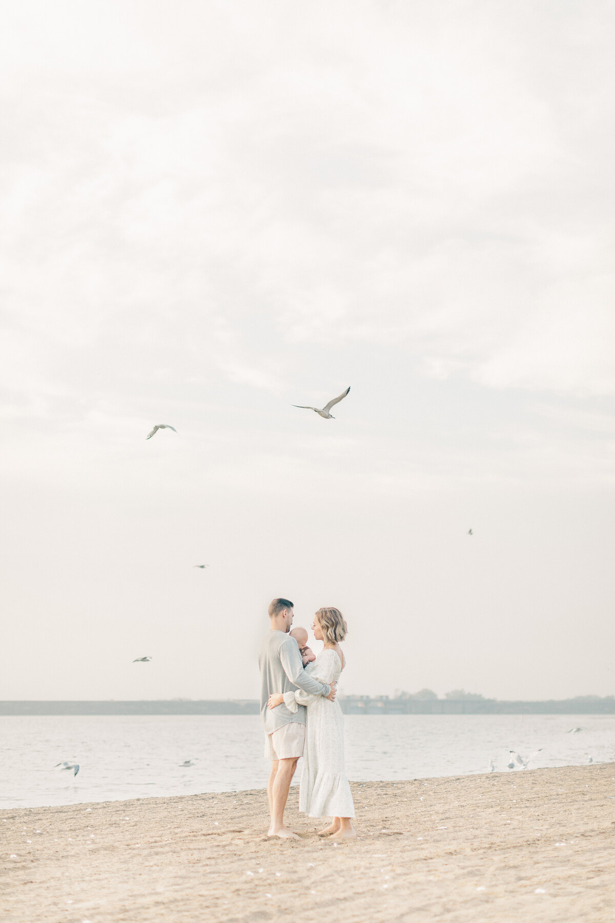Parents standing on the shore with their baby as birds fly overhead during a peaceful 6 month sitter milestone session at a Kansas City lake.