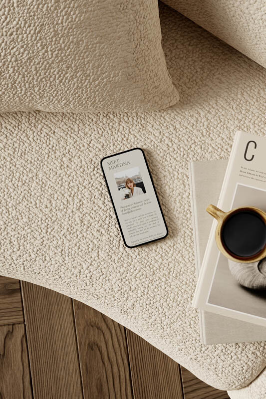 Flatlay of a mobile phone on a textured cream sofa, displaying a minimalist "Meet Martina" photographer bio page. Beside it rests a neutral-toned magazine and a cup of black coffee, creating a warm, editorial-inspired setting perfect for showcasing brand storytelling and intentional design.