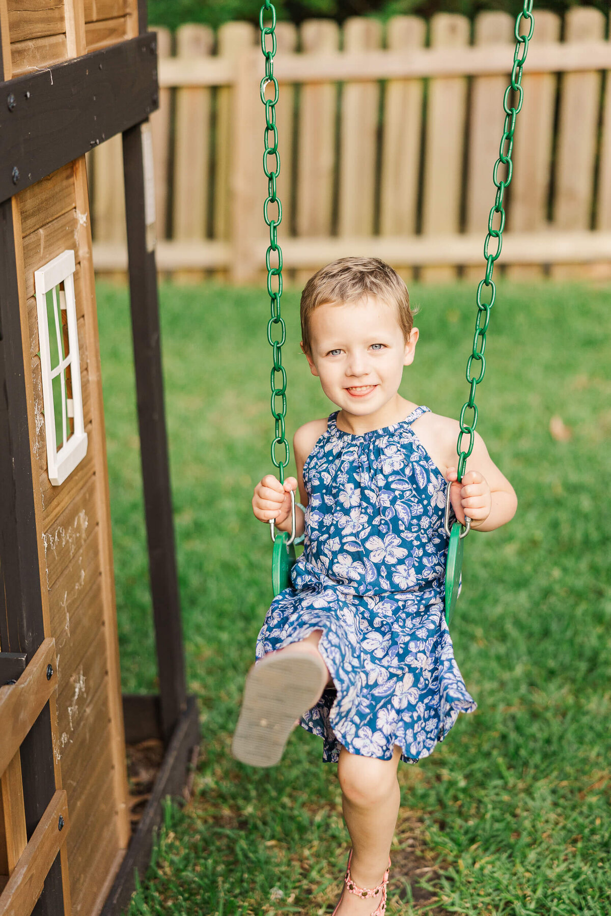 A child with short hair is smiling joyfully on a green swing in a backyard, captured beautifully by a Fayetteville NC family photographer. They wear a lovely blue and white floral dress. A wooden play structure sits to the left, complemented by a backdrop of a fence and lush grass.