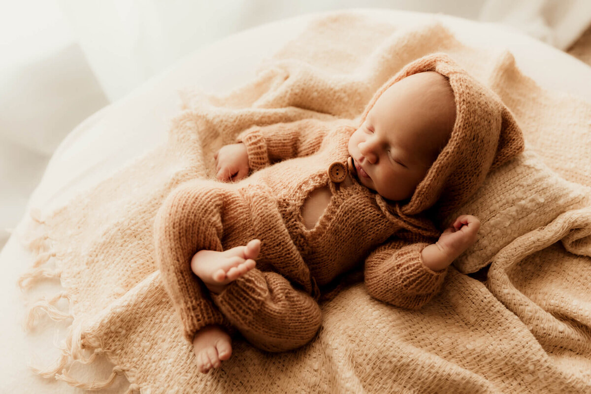 Baby boy wearing a brown hooded romper sleeps on a cream pillow and blanket near a window in OKC.
