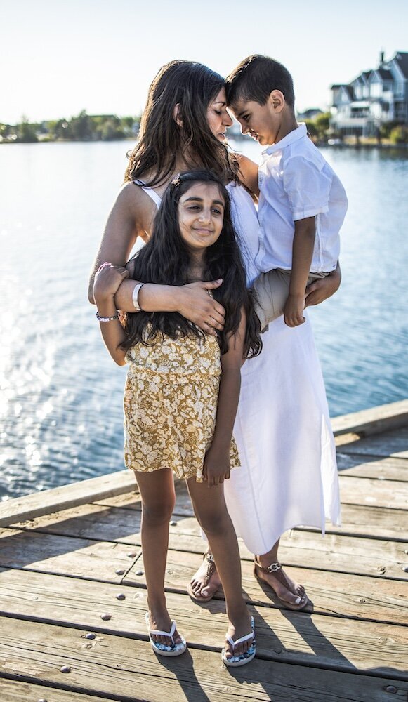 A mom in a white dress is holding her son on her hip and hugging her daughter with her other hand