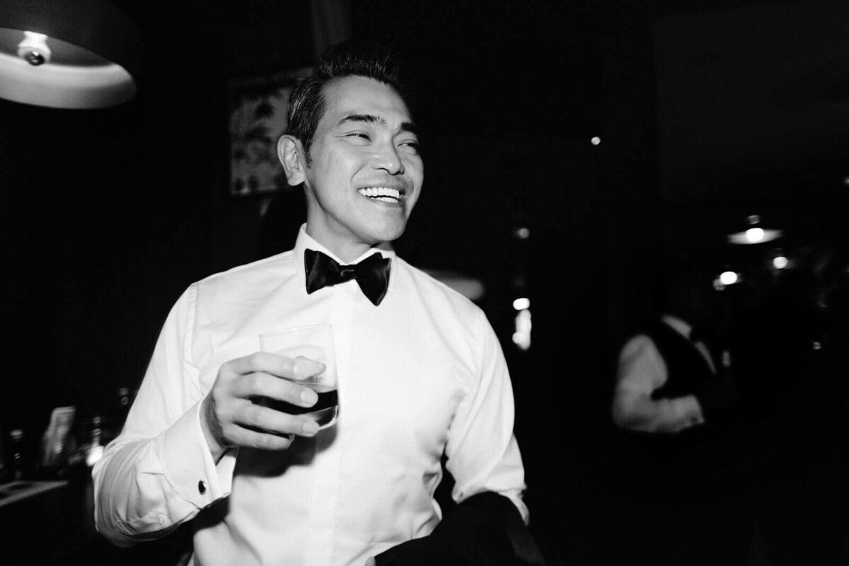 The groom is smiling, holding a glass of drink in The Skylark, New York. Wedding Image by Jenny Fu Studio