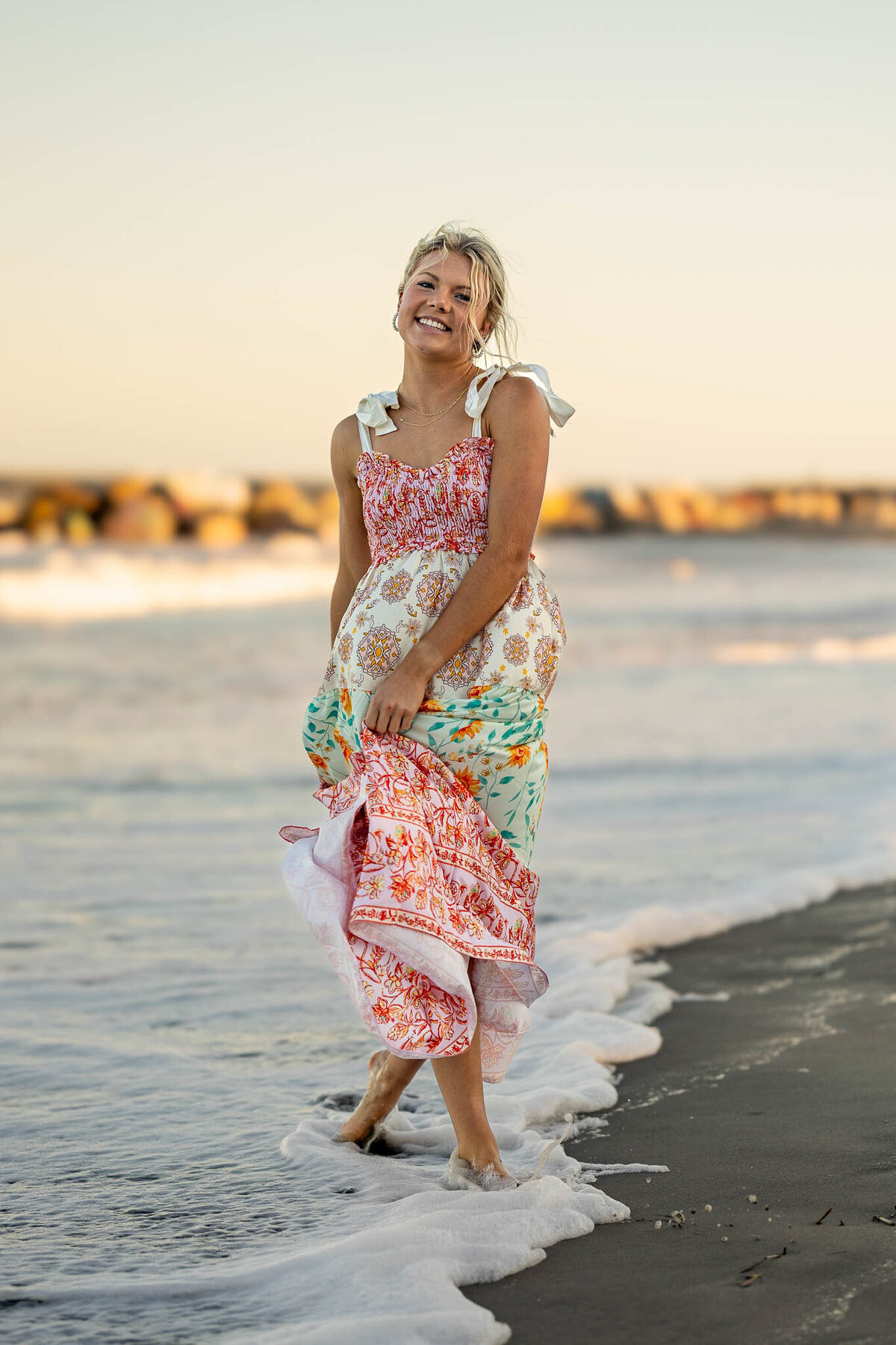 A senior girl in a floral maxi dress walks along the beach shoreline, with soft ocean waves and a pastel sky creating a dreamy coastal portrait.
