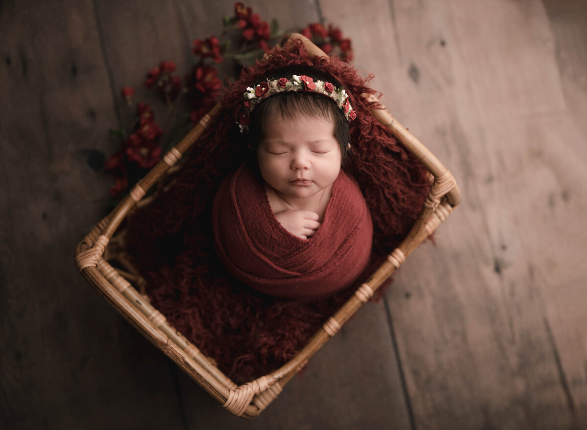 Baby girl is posed in a basket for her newborn photoshoot with Lake Elsinore's best newborn photographer Bonny Lynn Photography. Baby is swaddled in rust and her fingers are peeking out of the swaddle. Baby is wearing a matching floral headband.