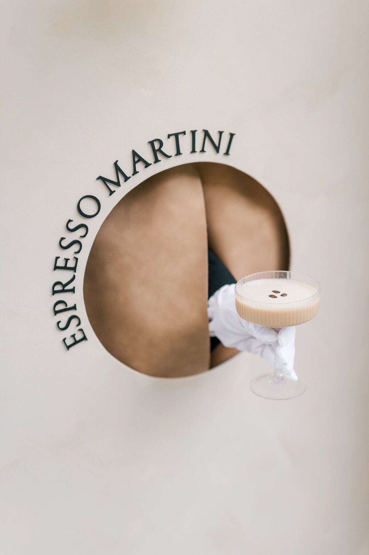 A hand holding an espresso martini out of a circular opening in a seating arrangement. 
