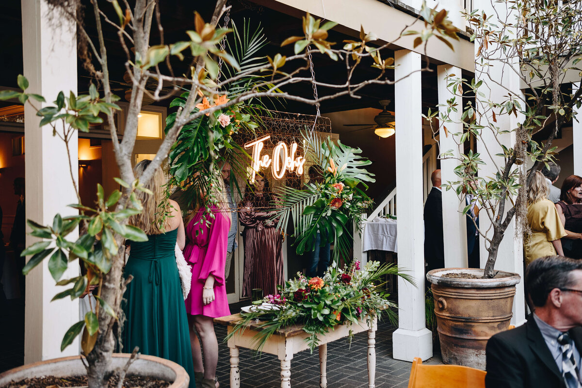Wedding guests gather near a neon sign reading The Oaks surrounded by lush greenery and floral arrangements in a courtyard with potted trees and white pillars.