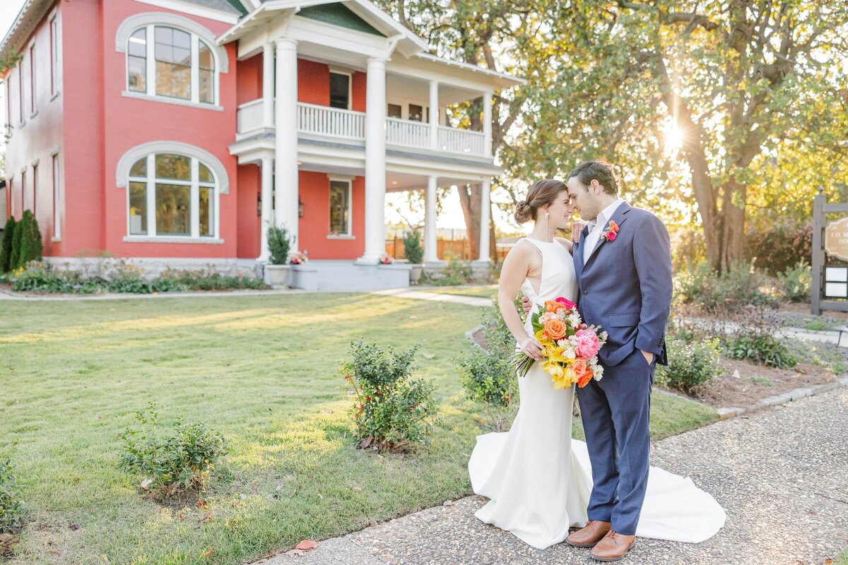 Newlyweds snuggle for a kiss while standing in the front garden of a pink wedding venue