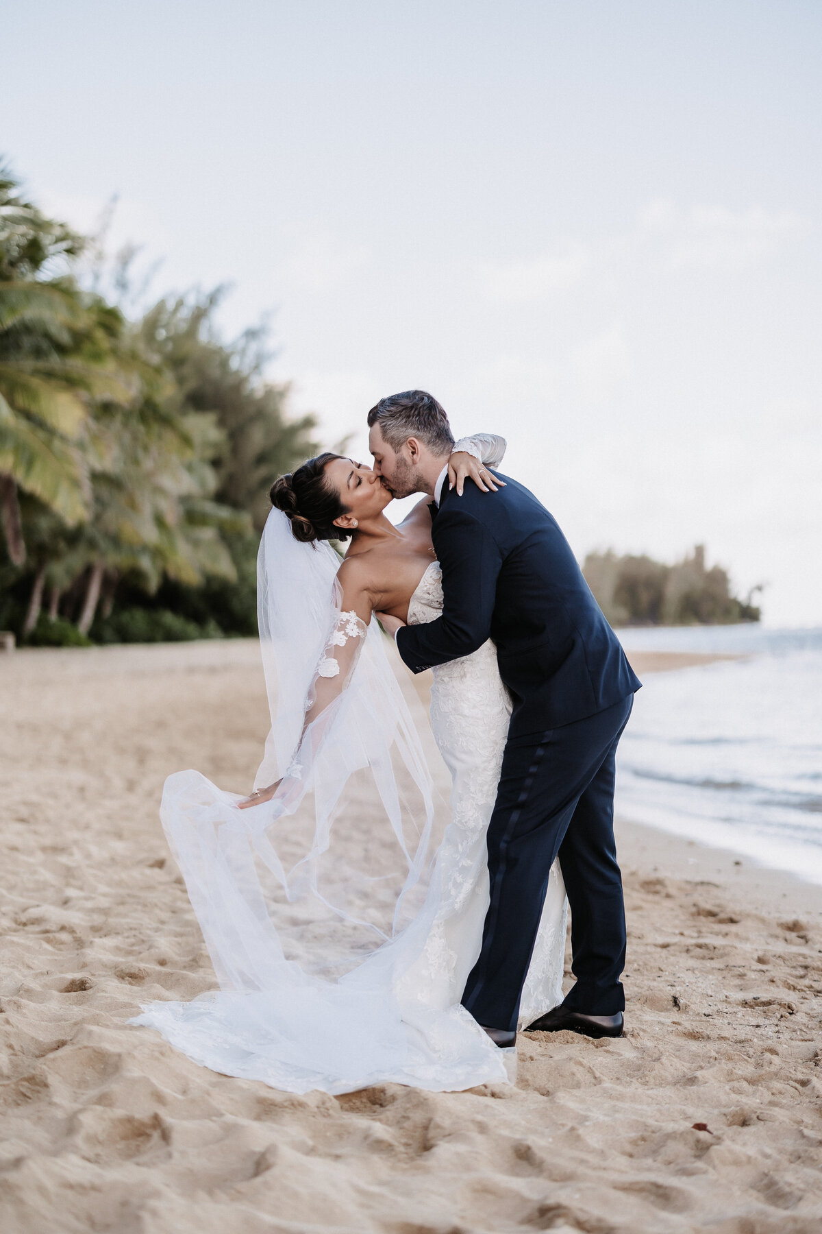 Bride and groom kissing on the beach dipped back, in front of palm trees